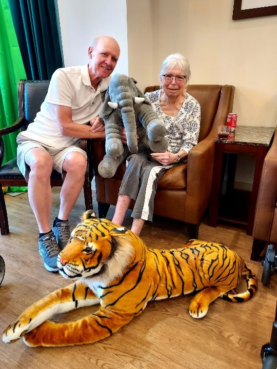An elderly woman and a man sitting indoors, holding a plush elephant and with a plush tiger on the floor in front of them.