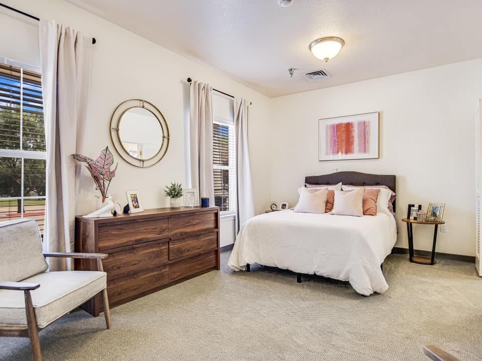 A cozy and well-decorated bedroom featuring a bed with decorative pillows, a wooden dresser with a circular mirror above, and soft lighting.