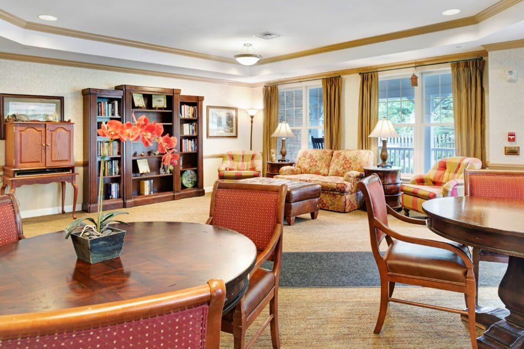 A cozy lounge area featuring comfortable seating, a wooden table with chairs, and a bookshelf in a well-lit room.