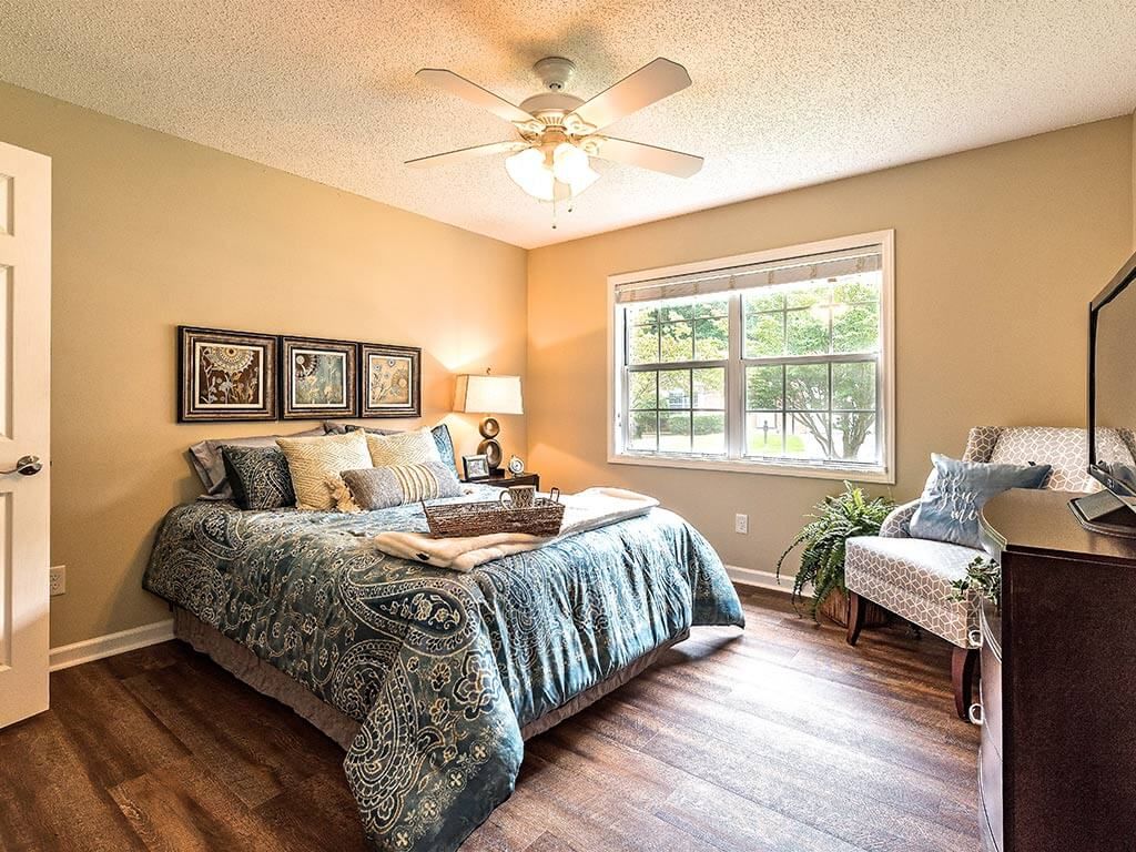 A cozy bedroom featuring a well-decorated bed, wooden flooring, and a large window allowing natural light to brighten the space.