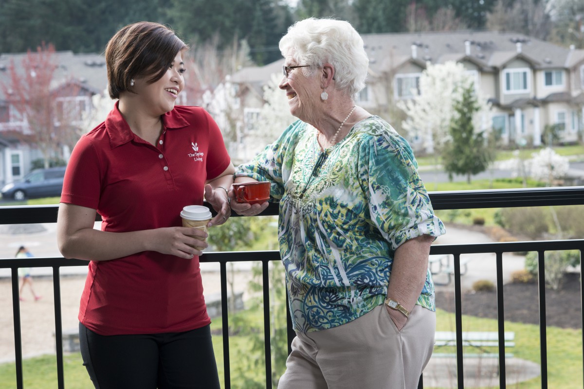 A senior woman and a younger woman enjoying a conversation on a balcony while holding coffee cups.