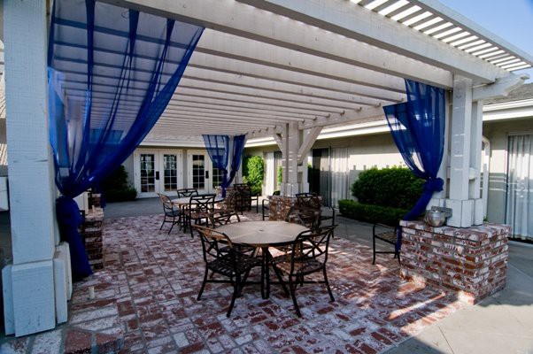 A charming outdoor patio area featuring tables and chairs under a pergola with blue curtains, set on a brick floor.
