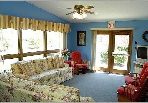 A cozy living room with floral sofas, an armchair, and a ceiling fan, illuminated by natural light through large windows.