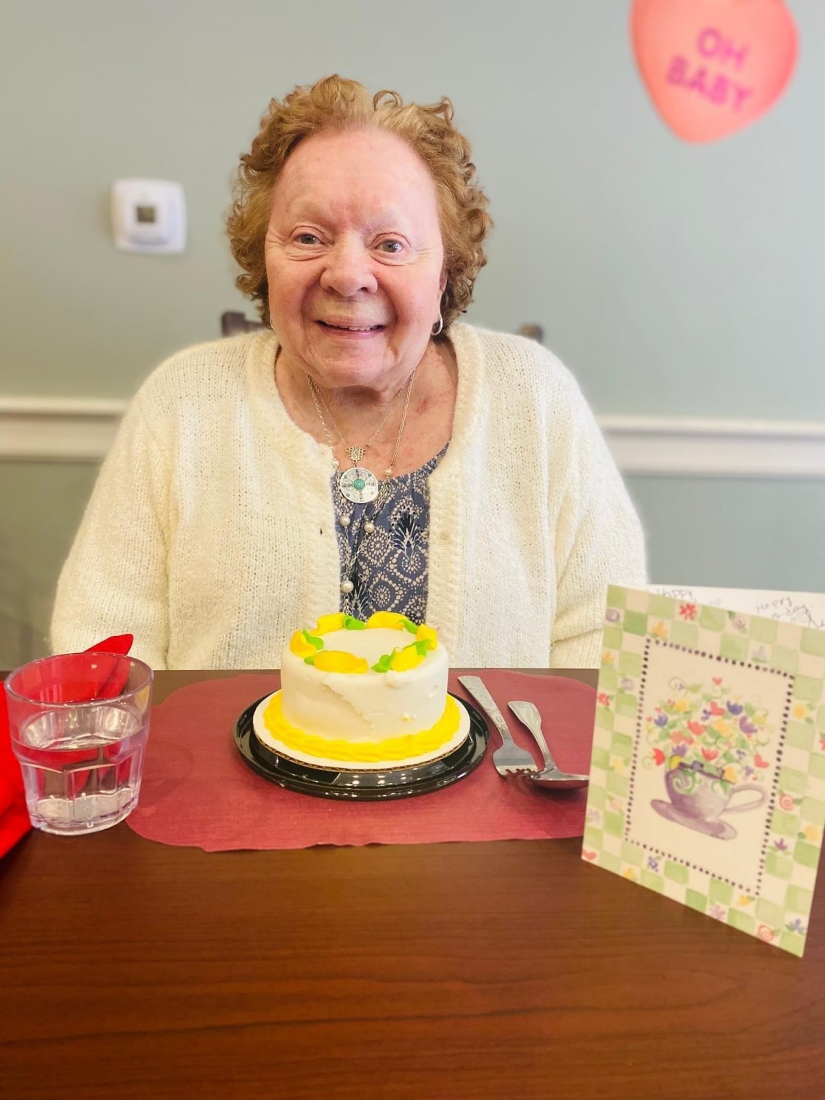 An elderly woman sitting at a table with a small birthday cake and a greeting card, smiling during a celebration.