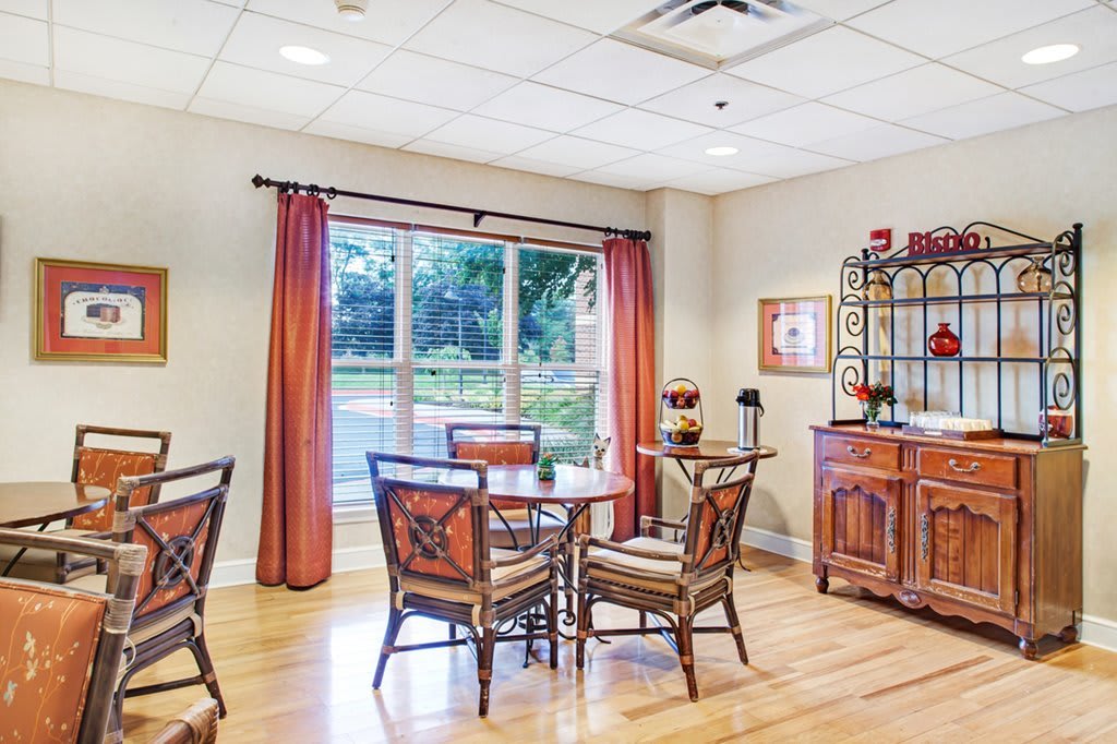 A cozy dining area with a table and chairs, featuring large windows with red curtains and a hardwood floor.