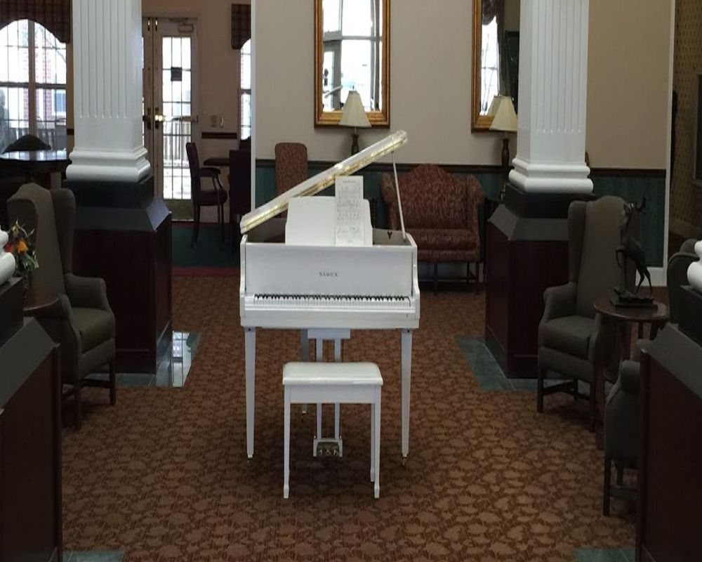 A white grand piano is placed in the center of a lobby with chairs, columns, and a patterned carpet.