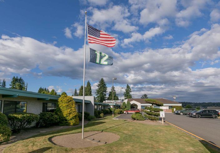 An American flag and a '12' flag fly outside a building on a sunny day with clouds in the sky.