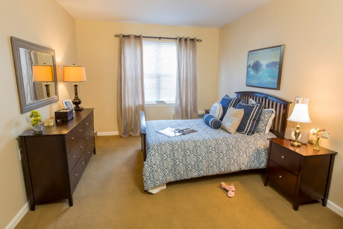 A cozy, well-decorated bedroom featuring a bed with patterned bedding, a dresser with a lamp, and sunlight filtering through the window curtains.