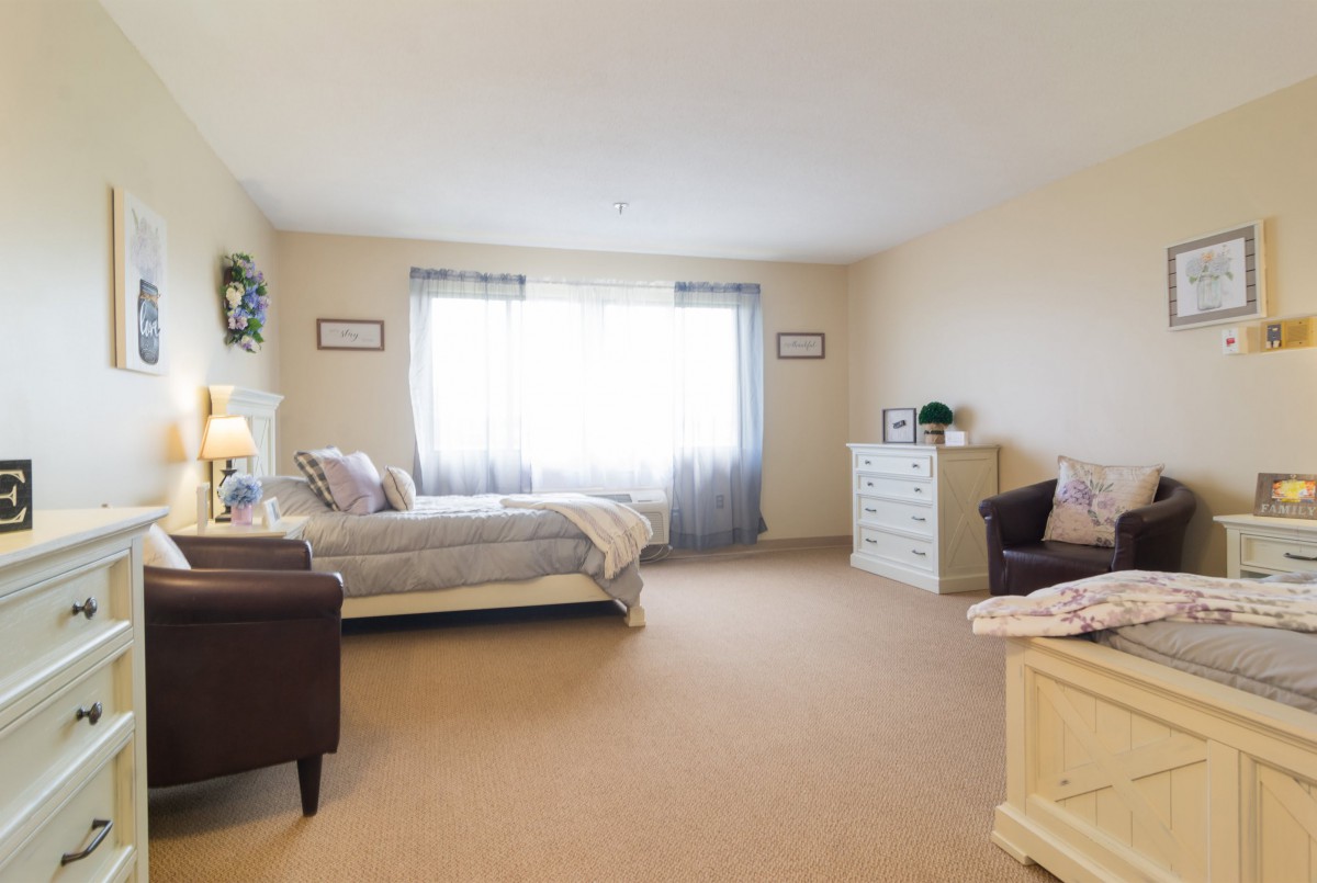 A spacious bedroom with neutral tones and sunlight streaming through the window, featuring a bed, dresser, and armchair.