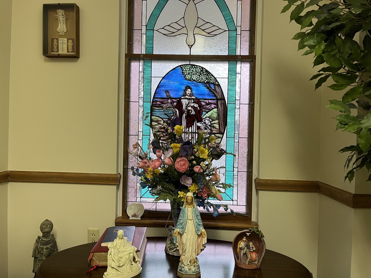 A decorative setup with religious statues and flowers in front of a stained glass window.