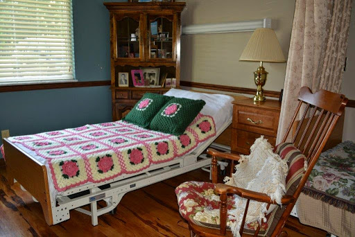 A cozy bedroom featuring a hospital bed adorned with a colorful quilt and decorative pillows, alongside an armchair and wooden furniture.