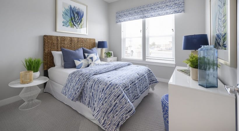A modern bedroom with a blue and white color scheme, featuring a bed adorned with patterned bedding and decorative pillows. The room is brightened by natural light from the large window, complemented by artwork on the wall.