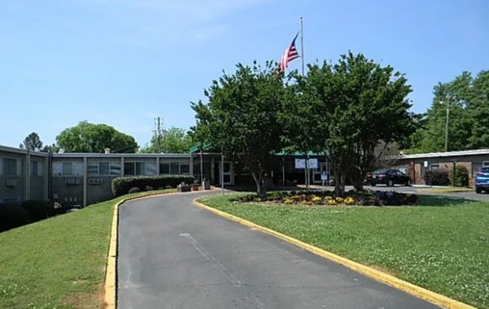 The image shows the entrance of a school building with a driveway, surrounded by trees and displaying an American flag on a flagpole.