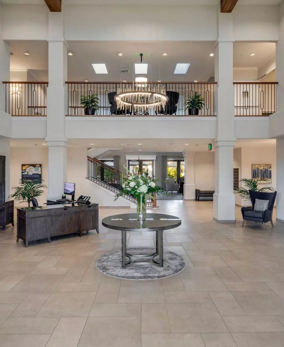 A spacious and elegantly designed lobby featuring modern furniture, a central table with flowers, and a grand chandelier.