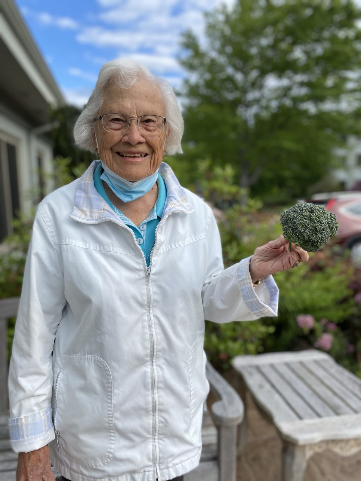 An elderly woman stands outdoors, smiling and holding an object in her hand.