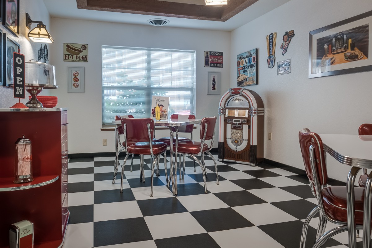 A vintage-style diner room with a black and white checkered floor, red chairs, and a classic jukebox.
