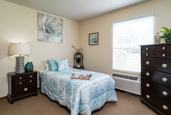 A cozy bedroom with a neatly made twin bed, featuring blue bedding and decorative pillows. The room is well-lit by a large window, and adorned with wall art and furniture.