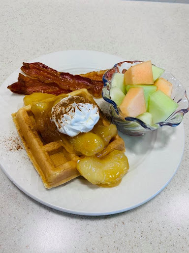 A delicious breakfast plate featuring waffles topped with peaches and whipped cream, crispy bacon, and a side of fresh melon.