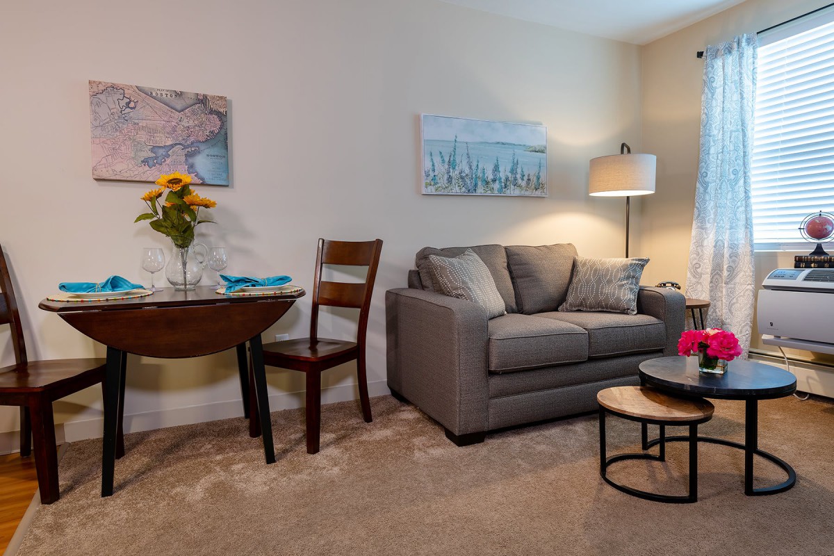A cozy living room with a gray sofa, wooden chair, and round coffee table, decorated with artwork and a standing lamp.
