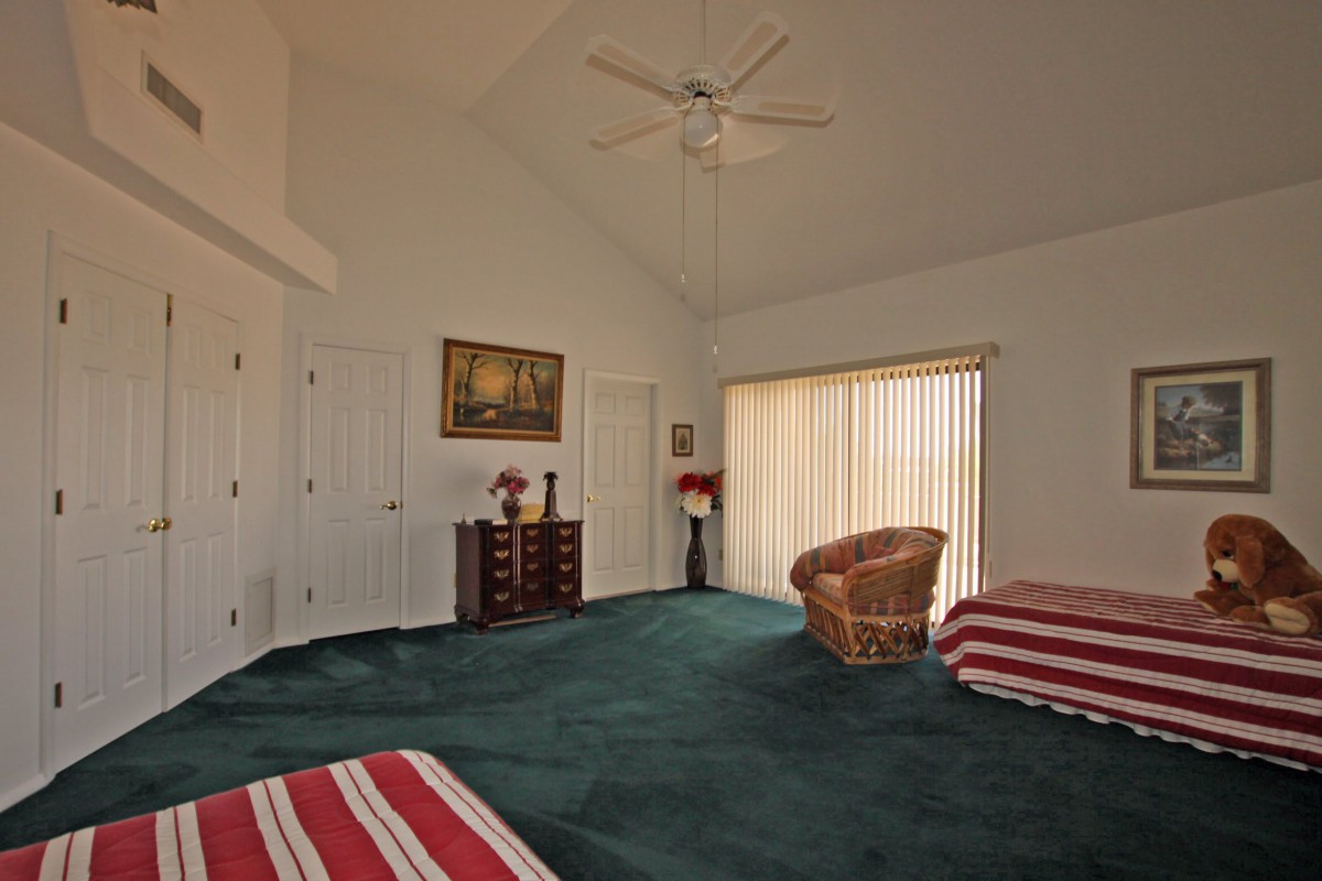 A spacious bedroom with green carpet, featuring a ceiling fan, wicker chair, dresser, and striped bedding.