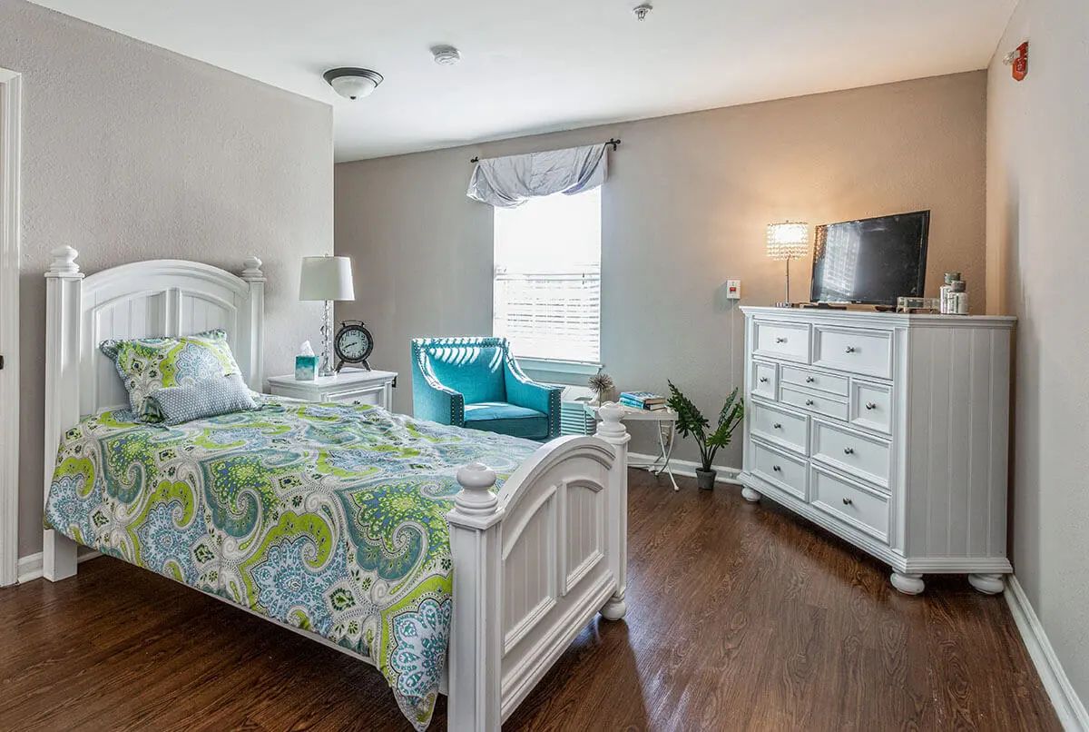 A cozy bedroom with a bed, dresser, and teal chair near the window.