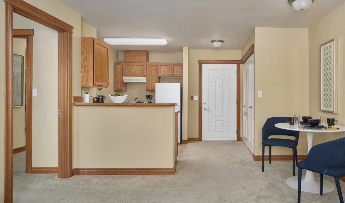 A cozy kitchen area in an apartment with wooden cabinets and a carpeted floor, featuring an open layout.