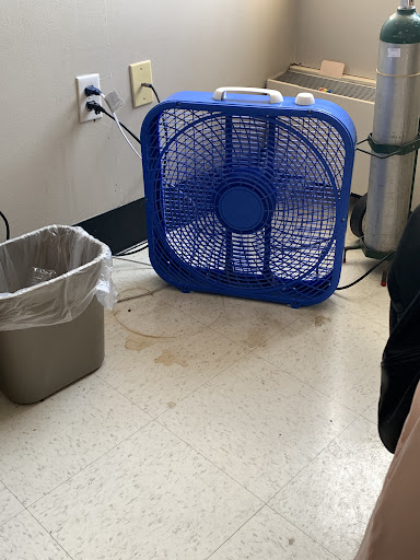 A blue fan placed next to a trash can in an office setting.