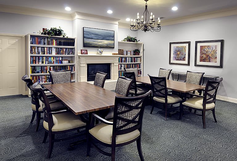 A well-organized library meeting room featuring a central table surrounded by chairs, bookshelves, a fireplace, and elegant lighting.
