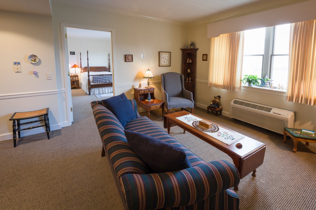 A cozy living room with a striped sofa, wooden coffee table, and armchair, leading into a bedroom.