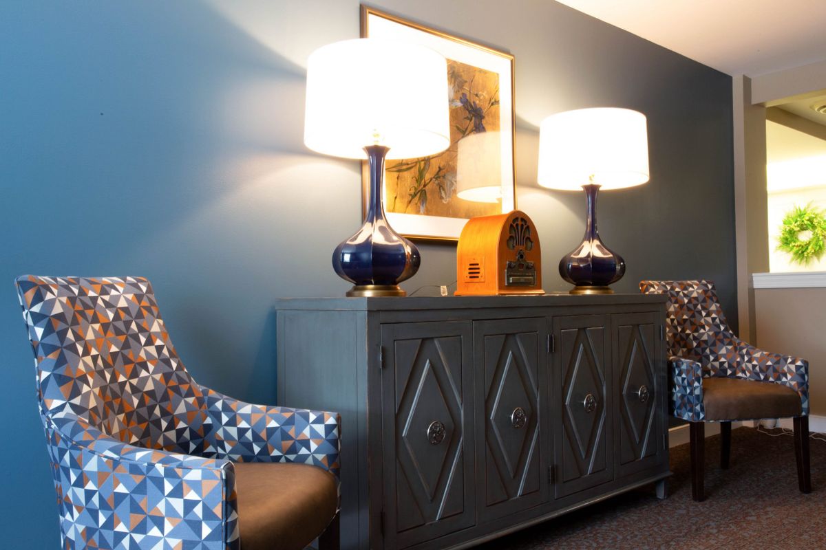 A stylishly decorated living room features a dark cabinet flanked by patterned chairs and topped with matching lamps, set against a blue wall with artwork.