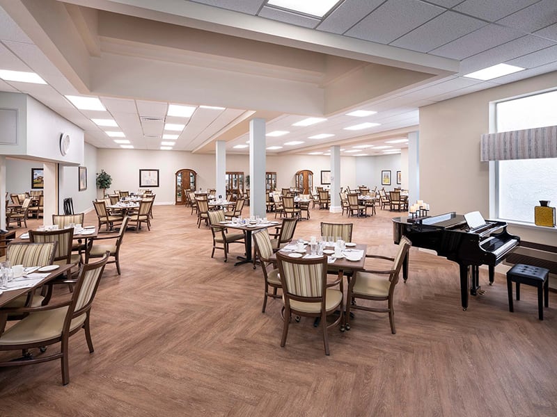 A spacious and empty dining hall with neatly arranged tables and chairs, featuring a piano in the corner.