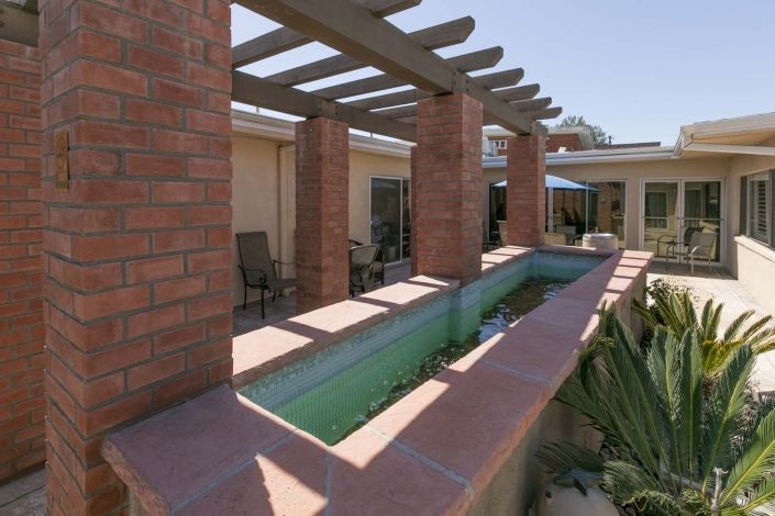 A patio area with a pergola supported by brick columns and an adjacent water feature on a sunny day.