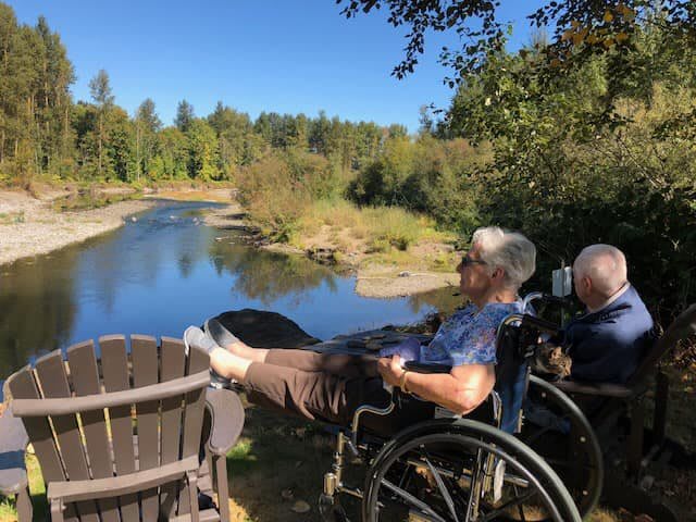 Two people in wheelchairs enjoy a peaceful view of the river and surrounding forest.