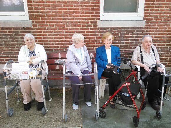 Four elderly women sitting on a bench with walkers in front of a brick wall.