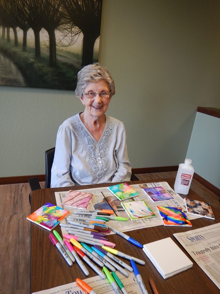 An elderly woman sitting at a table with art supplies, smiling warmly.