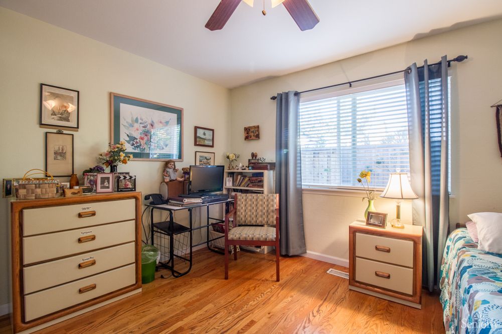A cozy home office setup with a desk, computer, and chair next to a window with curtains. The room features a wooden floor and is decorated with photo frames and a lamp.