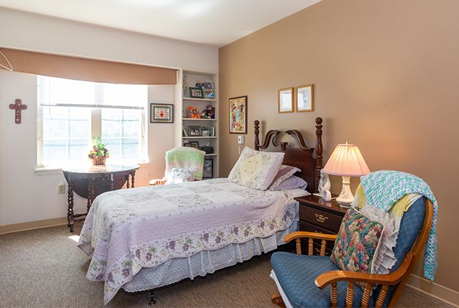 A cozy bedroom with a neatly made bed, a lamp on the nightstand, and a bookshelf beside the window.