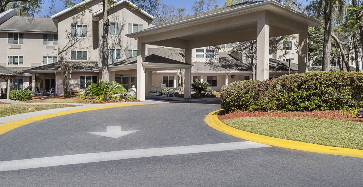 The image shows the entrance and driveway of a residential building, surrounded by landscaped gardens and trees.