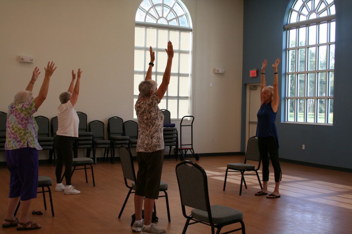 A group of women participating in a yoga class with chairs in a sunlit room.