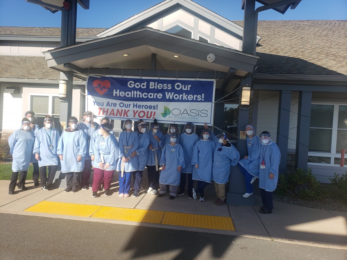 A group of healthcare workers standing in front of a building with a 'God Bless Our Healthcare Workers' banner.