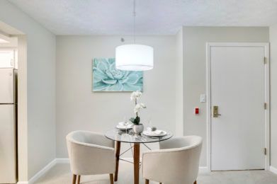 A modern dining area featuring a small circular glass table with two upholstered chairs, illuminated by a hanging light. A piece of abstract artwork adorns the wall.