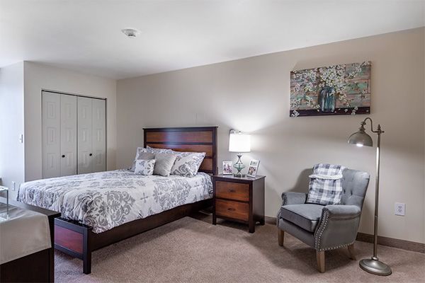 A cozy bedroom featuring a bed with patterned bedding, a wooden nightstand with a lamp, an armchair, and wall art.