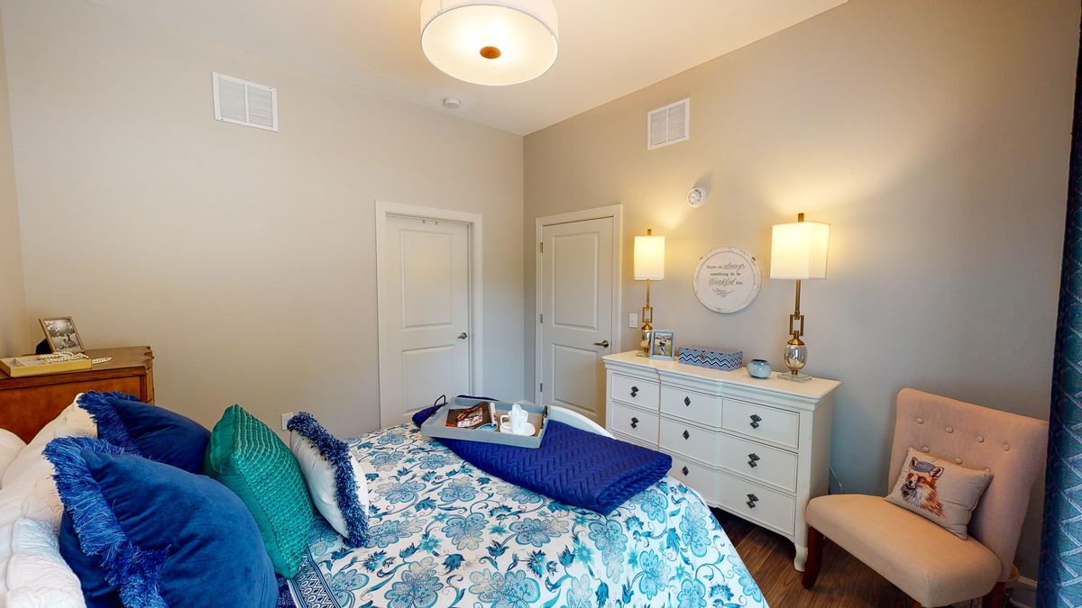 A cozy and modern bedroom with elegant decor, featuring a bed with patterned bedding, a white dresser with two lamps, and warm lighting.