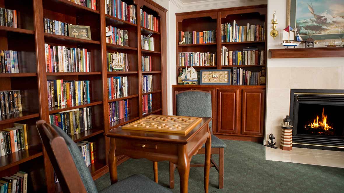 A cozy library room featuring wooden bookshelves filled with books and a chessboard on a table surrounded by chairs.