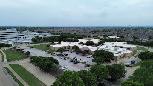 An aerial view of a school surrounded by green trees and a parking lot in a suburban setting.