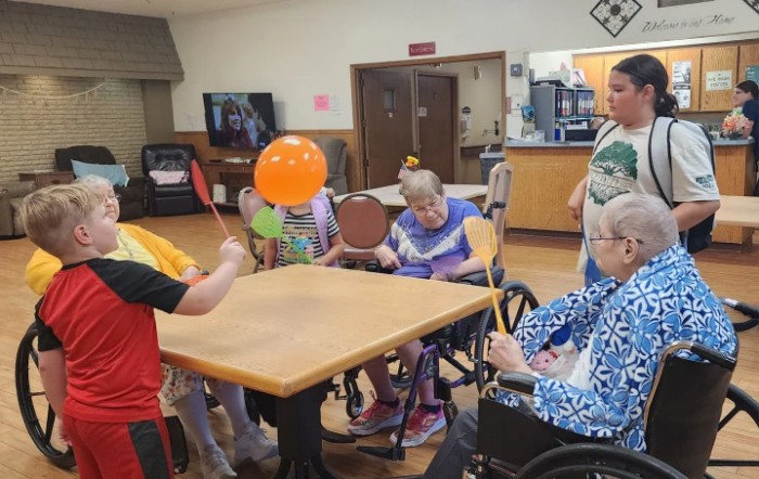 A group of elderly individuals engaging in a balloon activity indoors, seated around a table with some in wheelchairs.