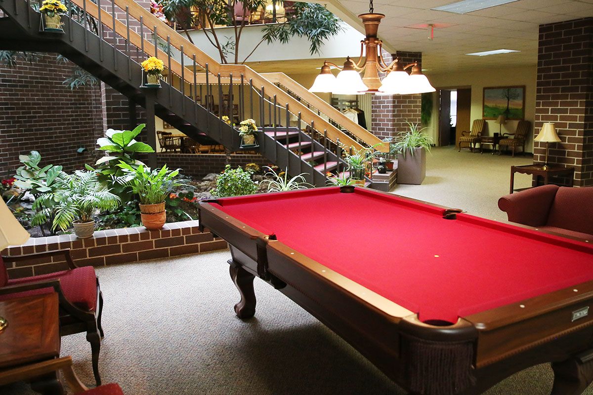 A cozy indoor lounge area featuring a red-felt pool table, surrounded by greenery and ambient lighting.