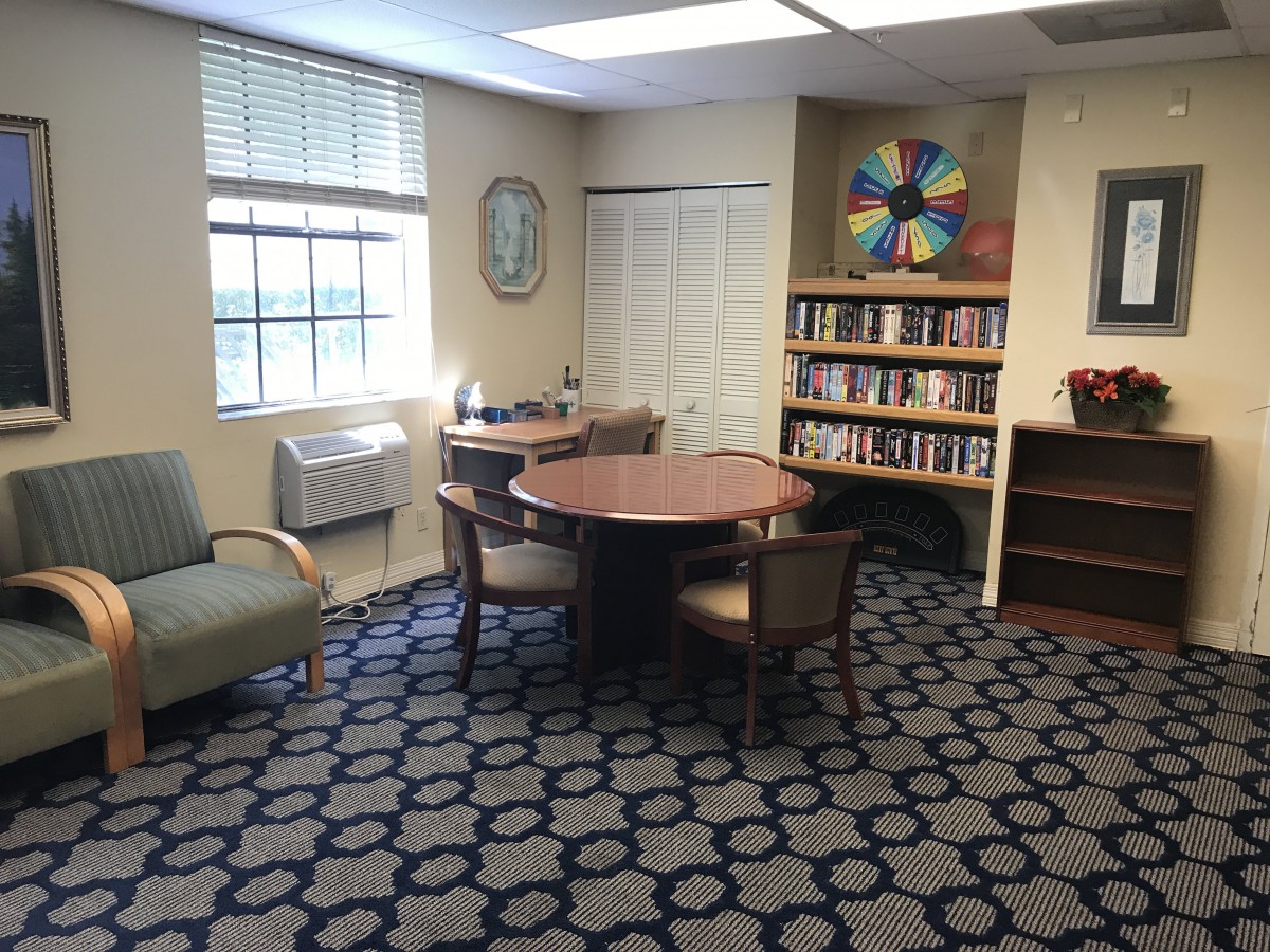 A cozy library corner with a round table, chairs, and bookshelves under natural light from a window.