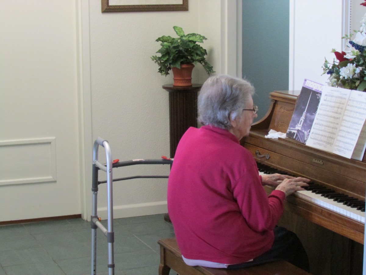 An elderly woman plays the piano with sheet music in an indoor setting, using a walker nearby.