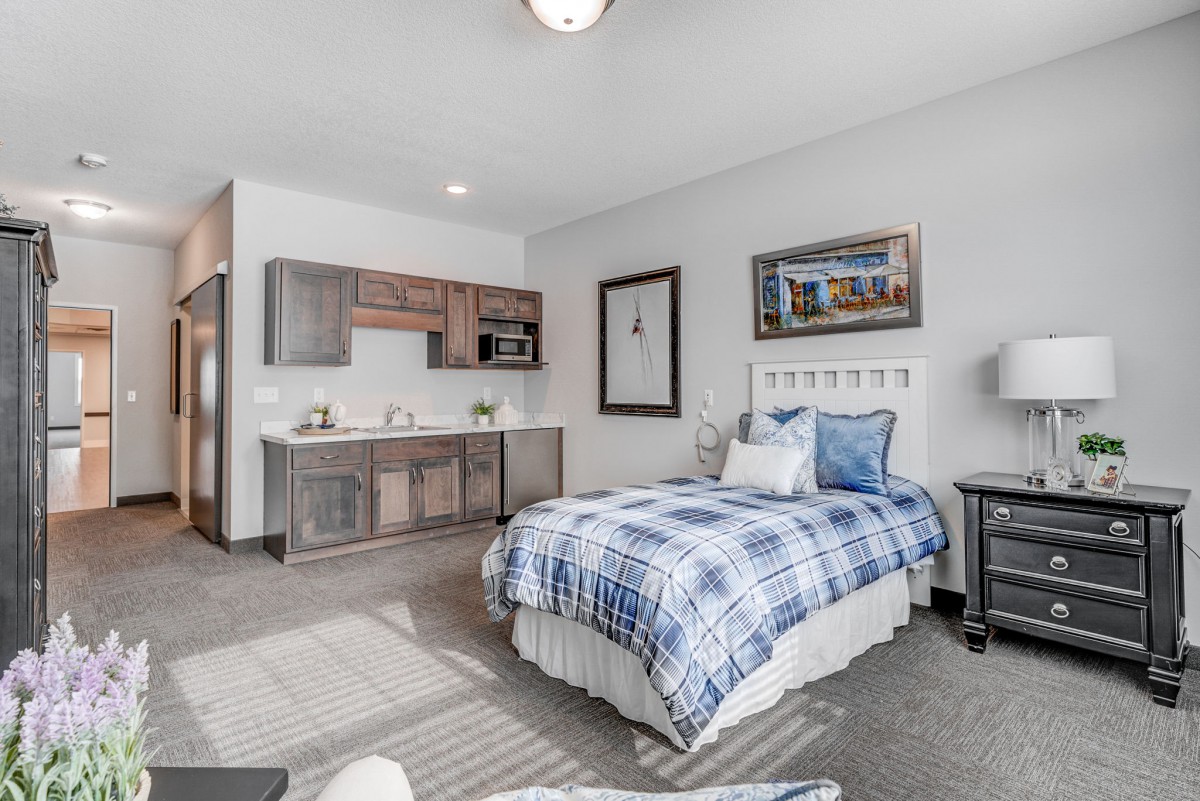 A modern bedroom with a single bed featuring blue plaid bedding, a kitchenette in the corner, and stylish wall art.